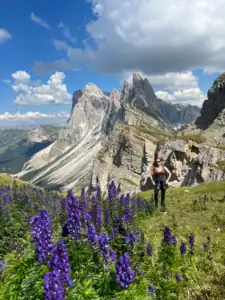 Me in the beautiful purple flowers and sunny skies with the massive Seceda ridgeline behind me.